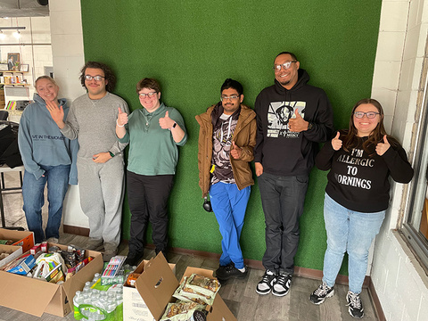 A group of people stand together near boxes of food, giving thumbs-ups.