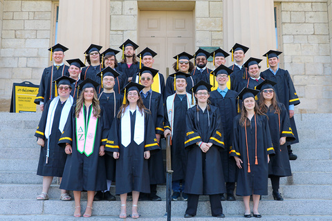 Graduating students stand in a group on the Old Capitol steps, wearing their caps and gowns.