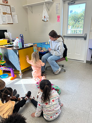 Caroline Keenan sits in a chair in front of a group of children, reading a picture book.