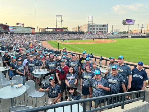 Dozens of ReConnect attendees stand together in a baseball stadium, wearing matching UI REACH shirts.