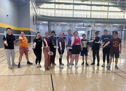 A group of students stand in front of a net, holding badminton rackets.