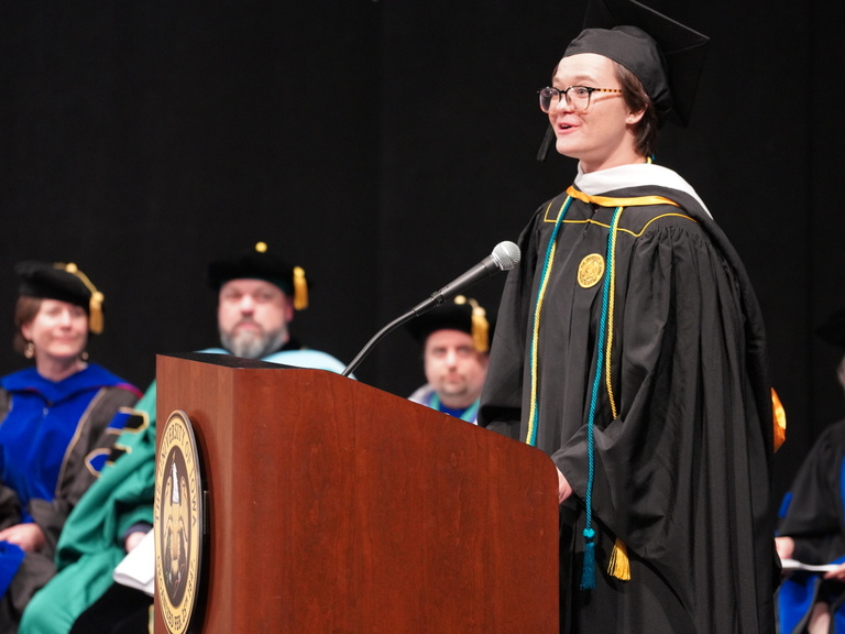 Samantha Pedek, Spring 2023 Student Commencement Speaker, stands at a podium, dressing in commencement garb, speaking to the audience.