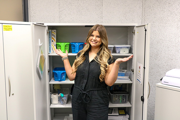 Abby stands in front of the Adaptive Equipment Closet with her hands raised.
