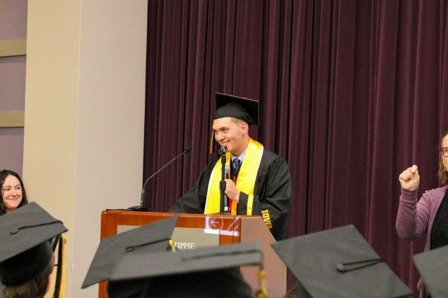 Simon Janelle stands at a podium, holding a microphone and wearing a cap and gown.