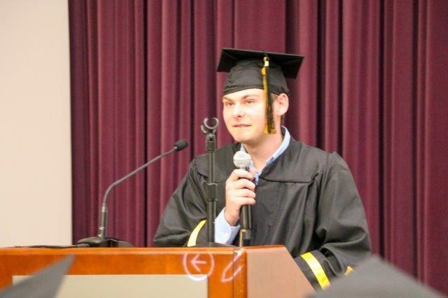 Eli Mogil stands at a podium, holding a microphone and wearing a cap and gown.