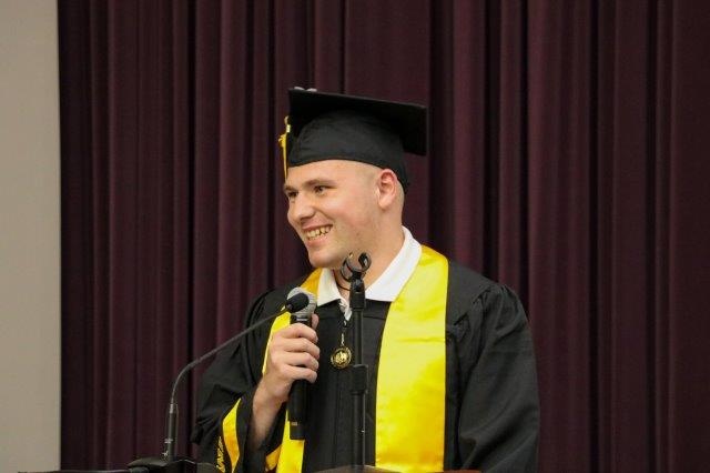 Tyler Mumm stands at a podium, holding a microphone and wearing a cap and gown.