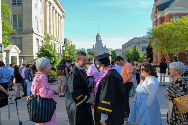 Two graduates in their caps and gowns shake hands outside of the Old Capitol Building, surrounded by family and friends.