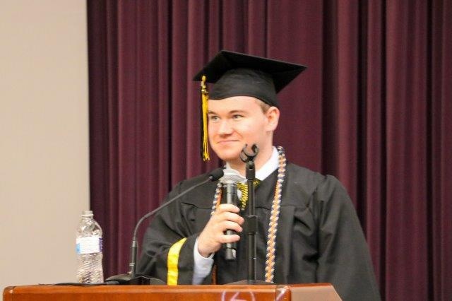 Mathew Springer stands at a podium, holding a microphone and wearing a cap and gown.