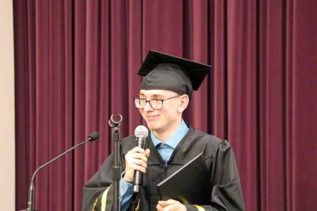 Ben Stangl stands at a podium, holding a microphone and wearing a cap and gown.