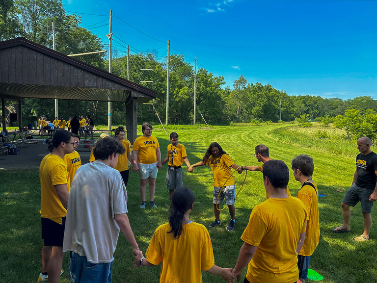A group of students stand in a circle outside, playing a game with a hula hoop.