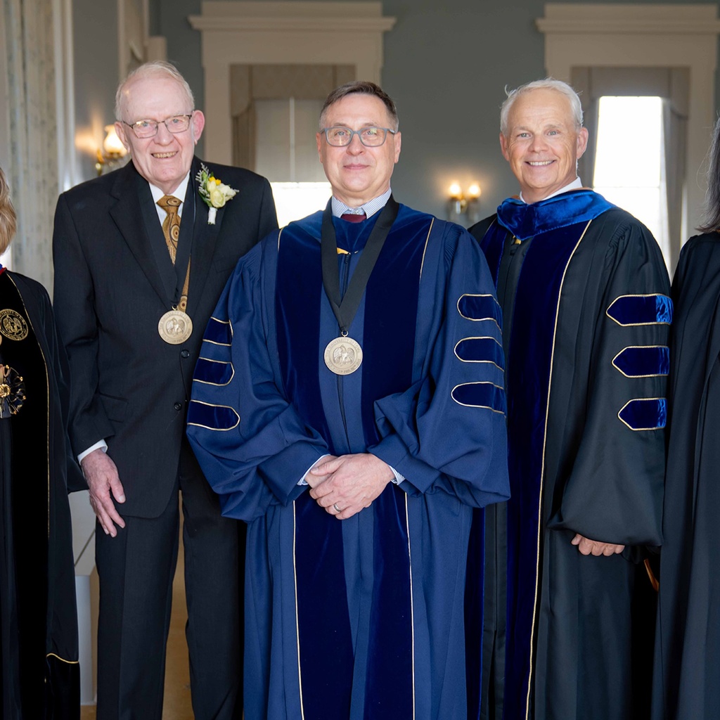 A professor wearing a ceremonial robe stands among a group of dignitaries, including UI President Barbara Wilson, during an investiture ceremony in the Old Capitol Senate Chambers.