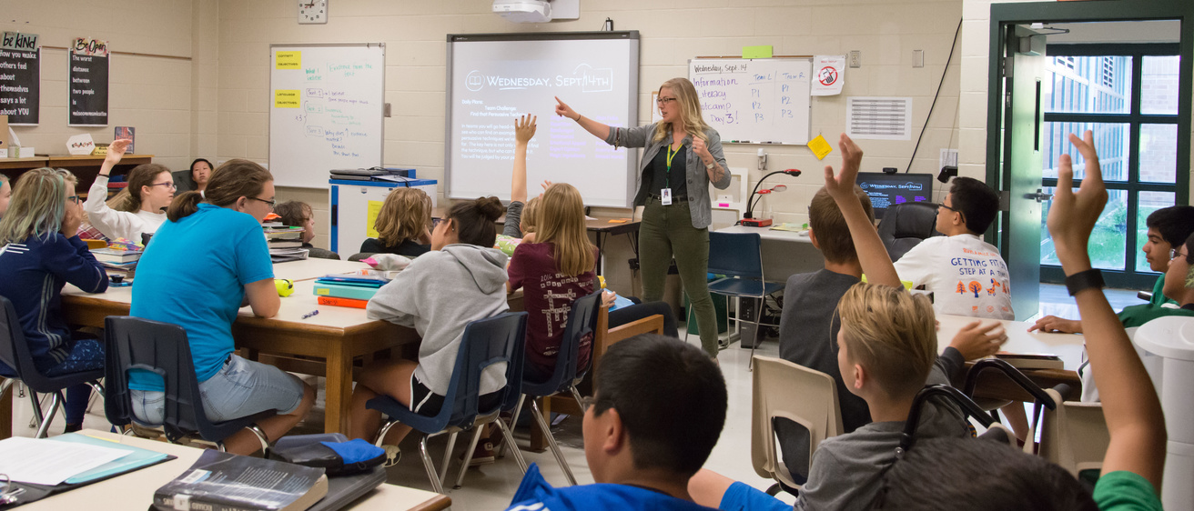 Teacher in middle school classroom with students raising hands.