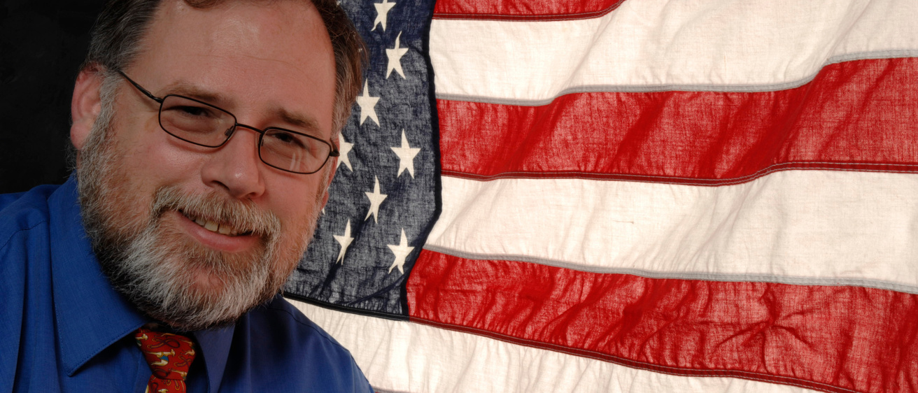Alumna and veteran John Mikelson in front of a U.S. flag