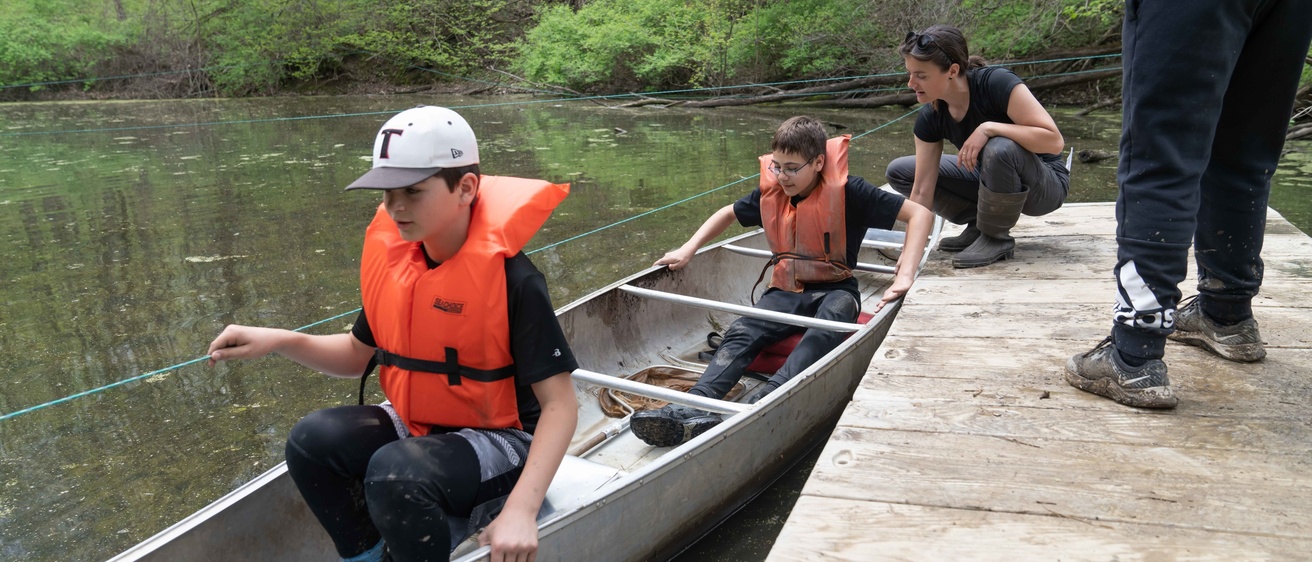 Campers wear life jackets and sit in a canoe on a pond.