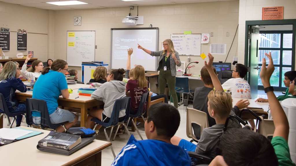 Teacher in middle school classroom with students raising hands.