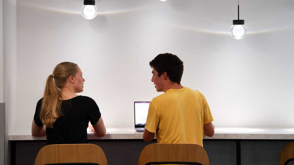 Two students sit next to each other looking at one another in front of a laptop computer