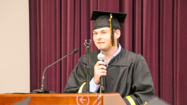 Eli Mogil stands at a podium, holding a microphone and wearing a cap and gown.